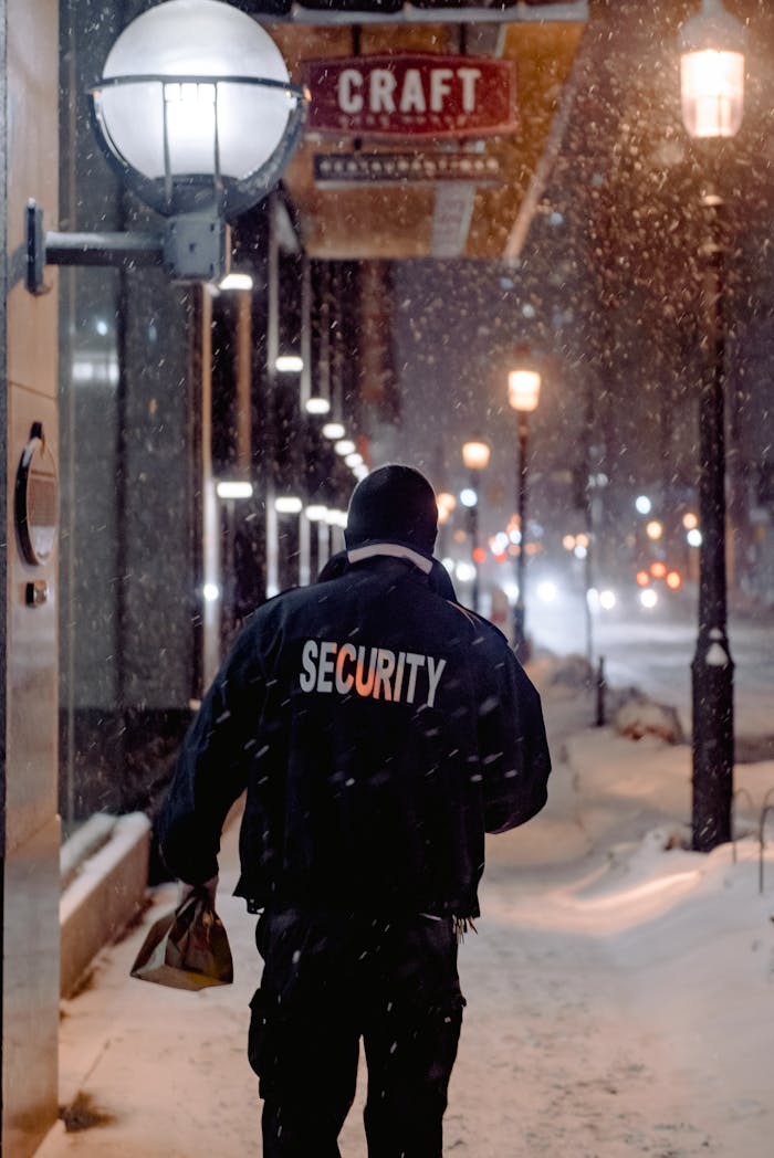 A back view of a security guard walking on a snowy street at night under street lights.